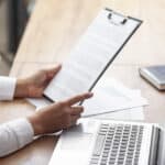 A person holding a contract pinned on a clipboard while in front of a laptop.