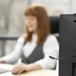 A woman working on her computer with another PC on her side, wearing a headset and looking at its monitor.