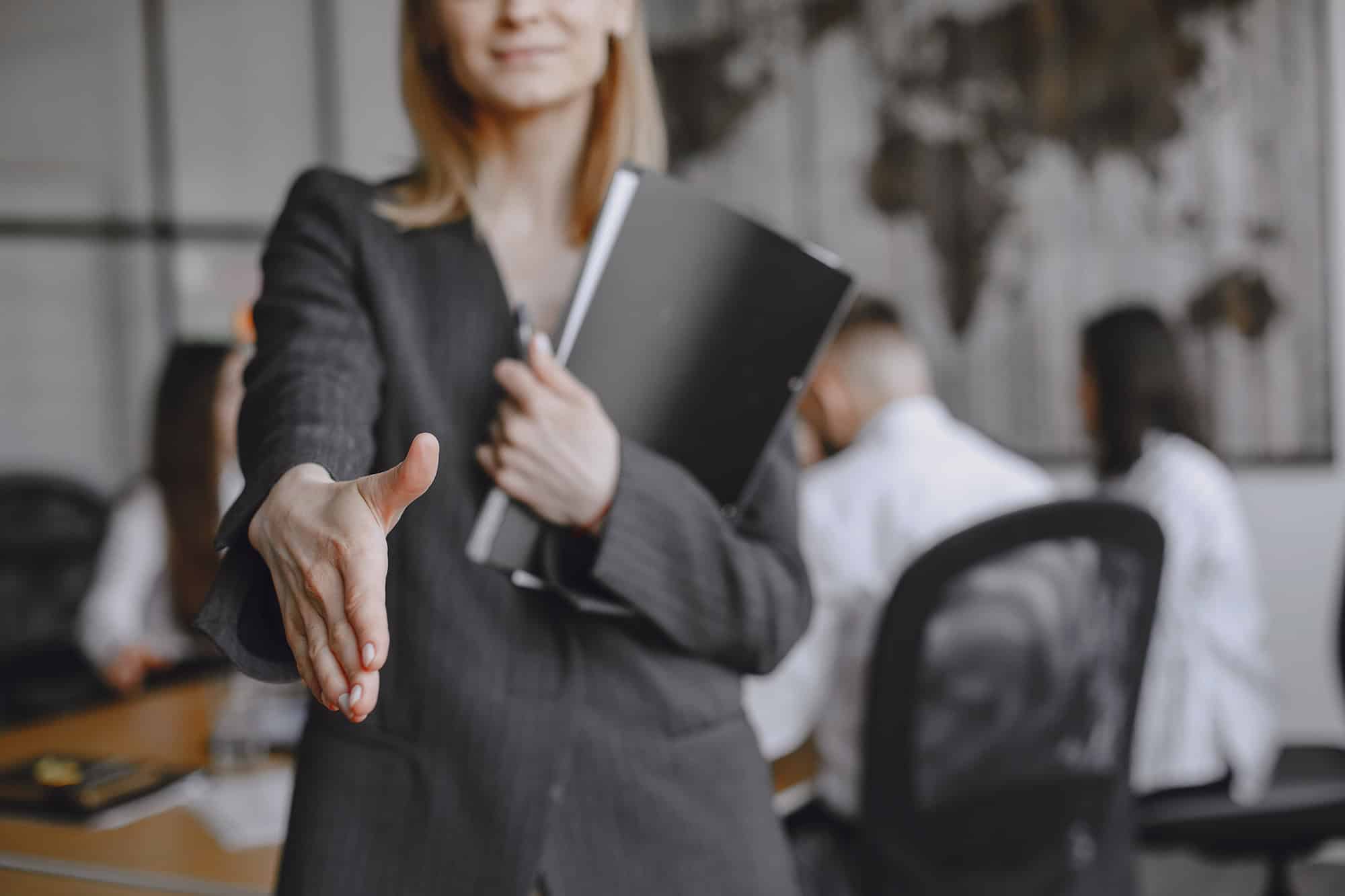 A woman wearing black striped corporate clothes is extending her hand out for a shake.