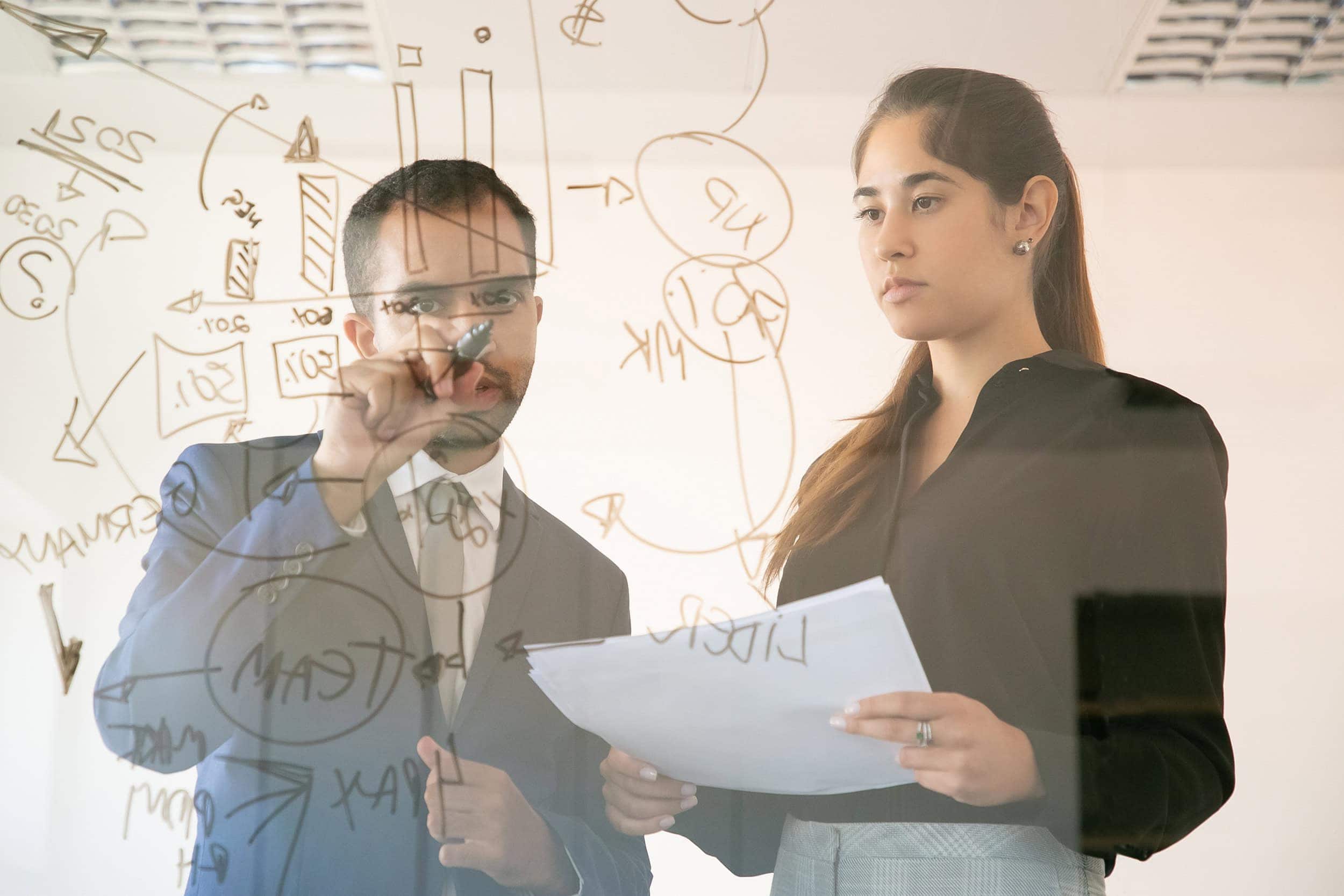 A man and a woman discussing work while scribbling a glass board.