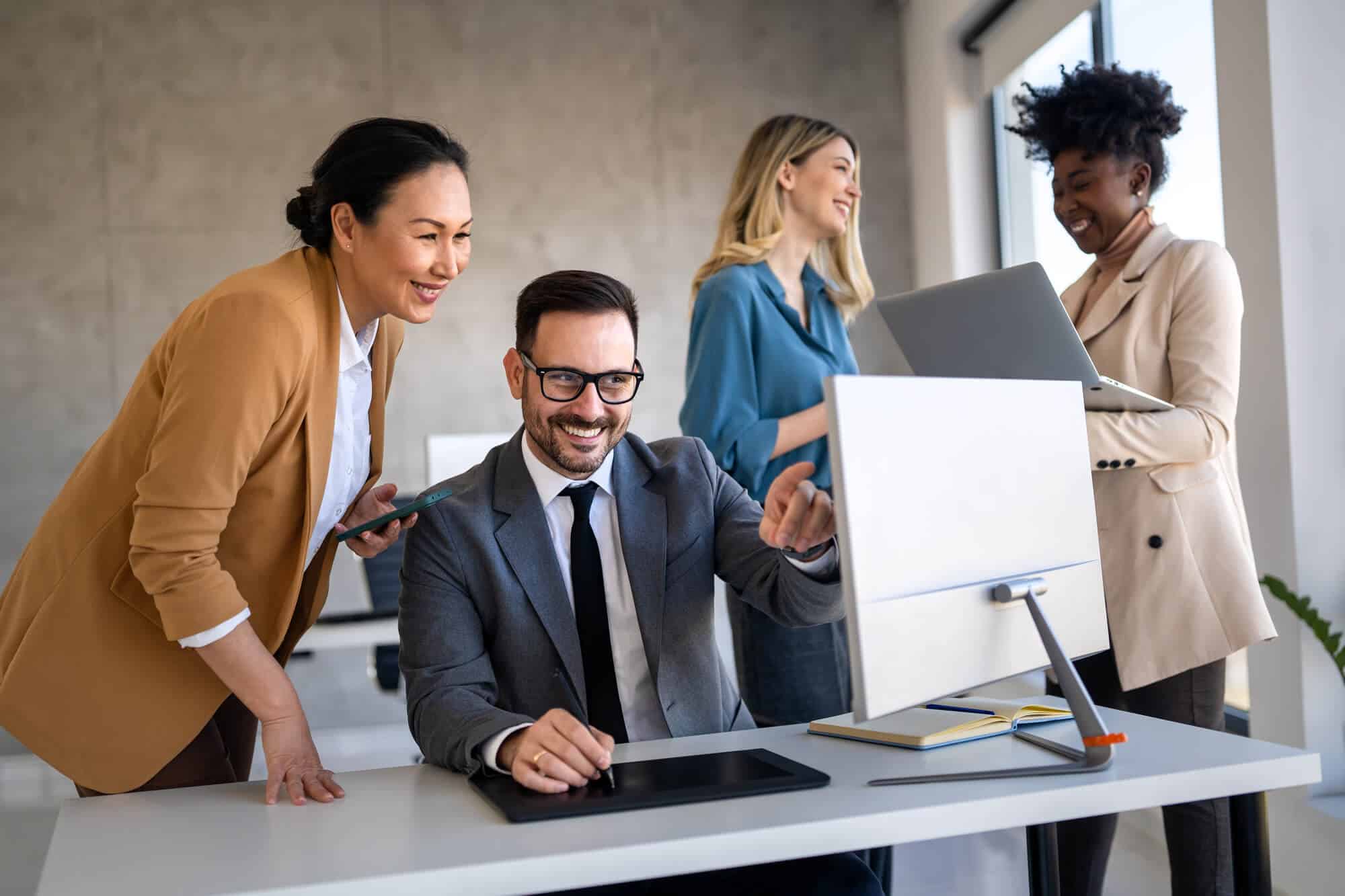 Four coworkers smile in the office. Two of them are looking at a desktop computer while the other two stand behind them.