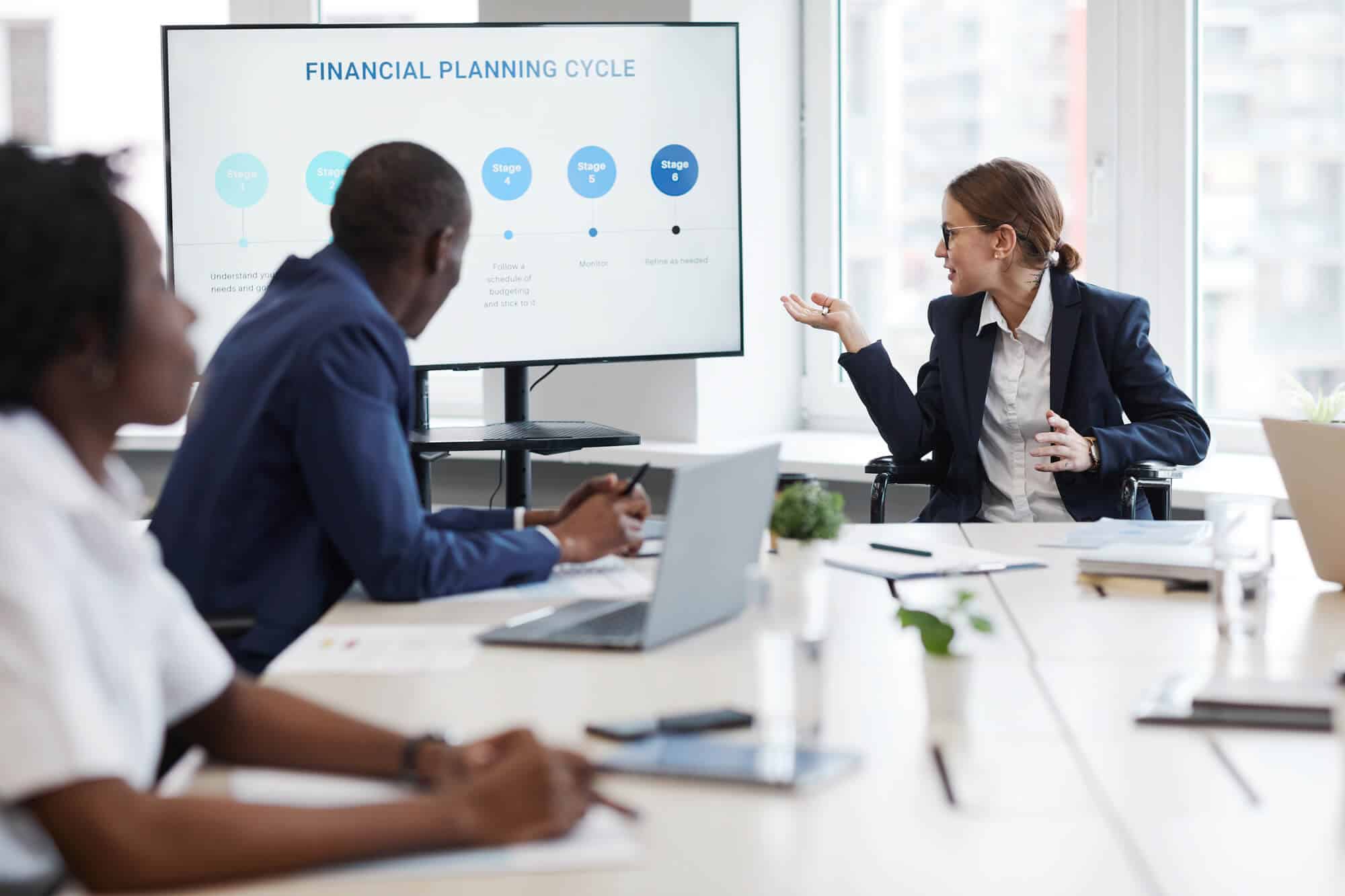 A woman gestures to a financial planning cycle presentation as her coworkers watch.