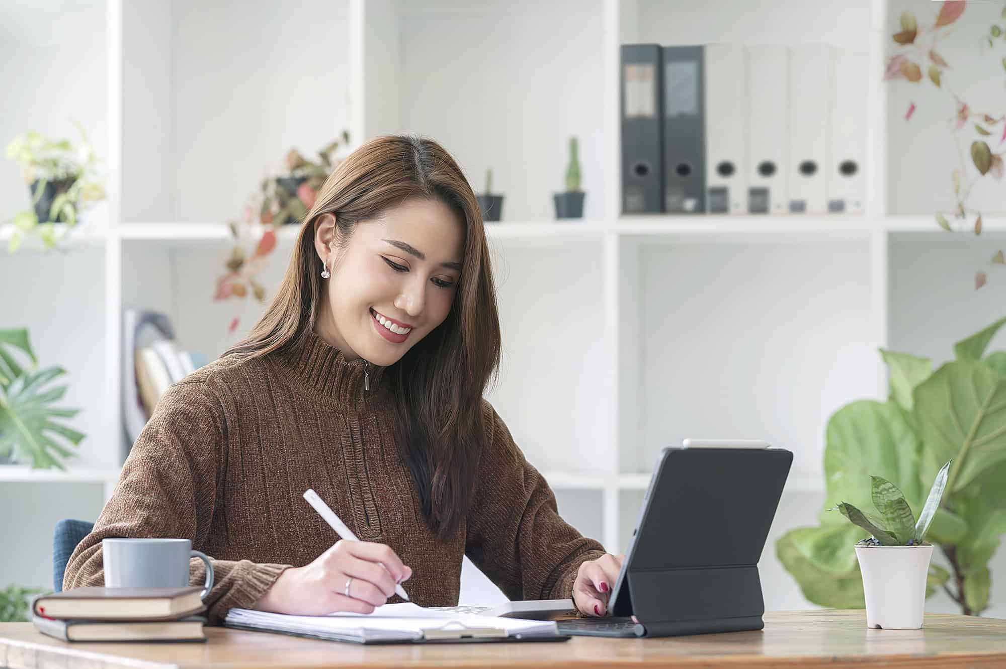 An HOA assistant writing notes while working on a laptop in a bright home office.