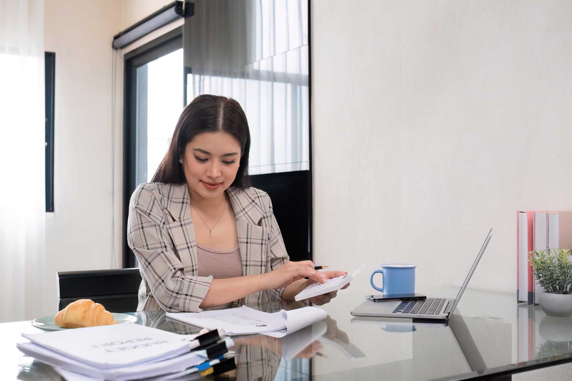 A woman wearing a blazer uses a calculator at her desk.
