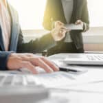 A co-worker is handing out a pile of paper to a man who is working at his desk.