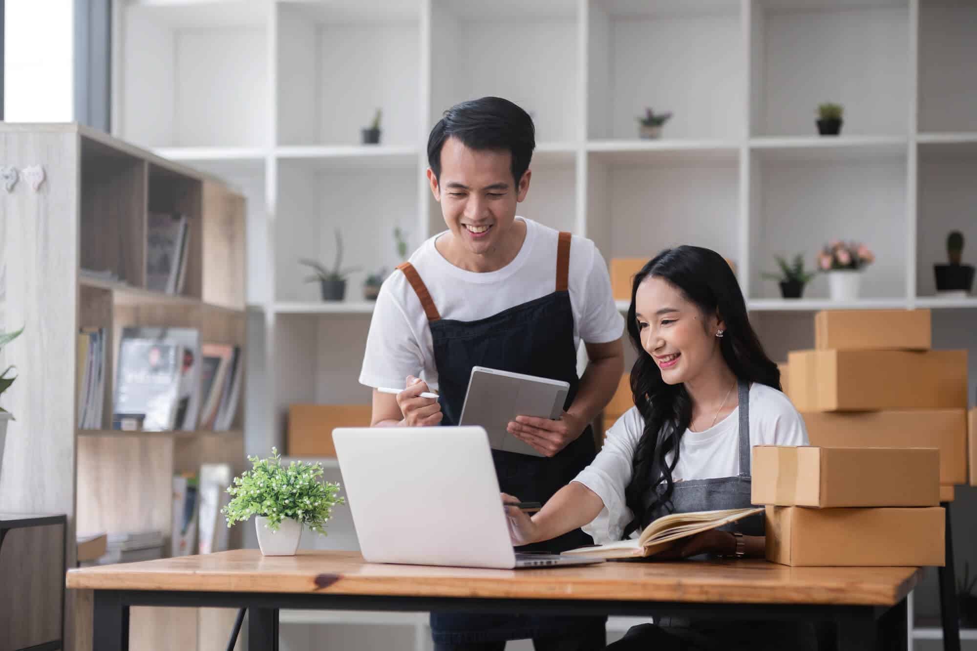 A man and a woman wearing aprons smile as they look at a laptop. They are surrounded by cardboard boxes.