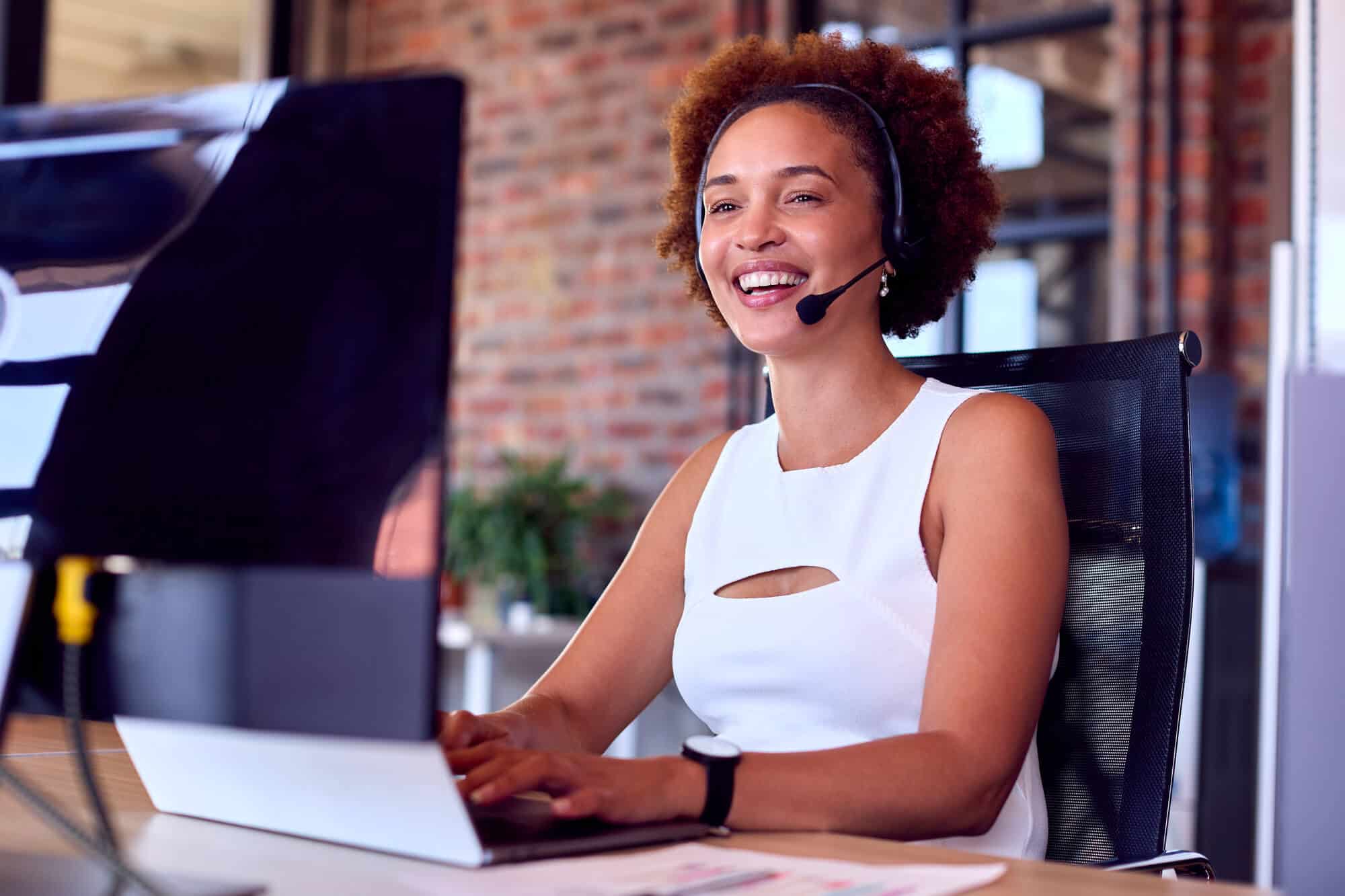 A woman wearing a white tank top and a black headset smiles as she looks at a computer.