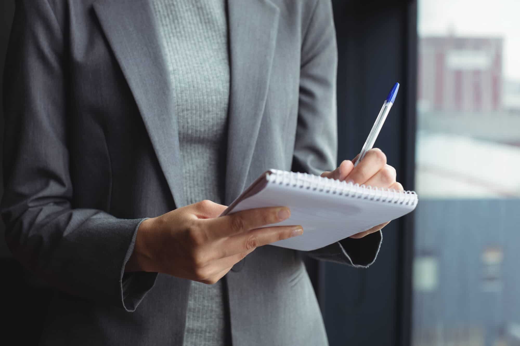 A close-up of a person in a gray sweater and gray blazer writing on a notepad.